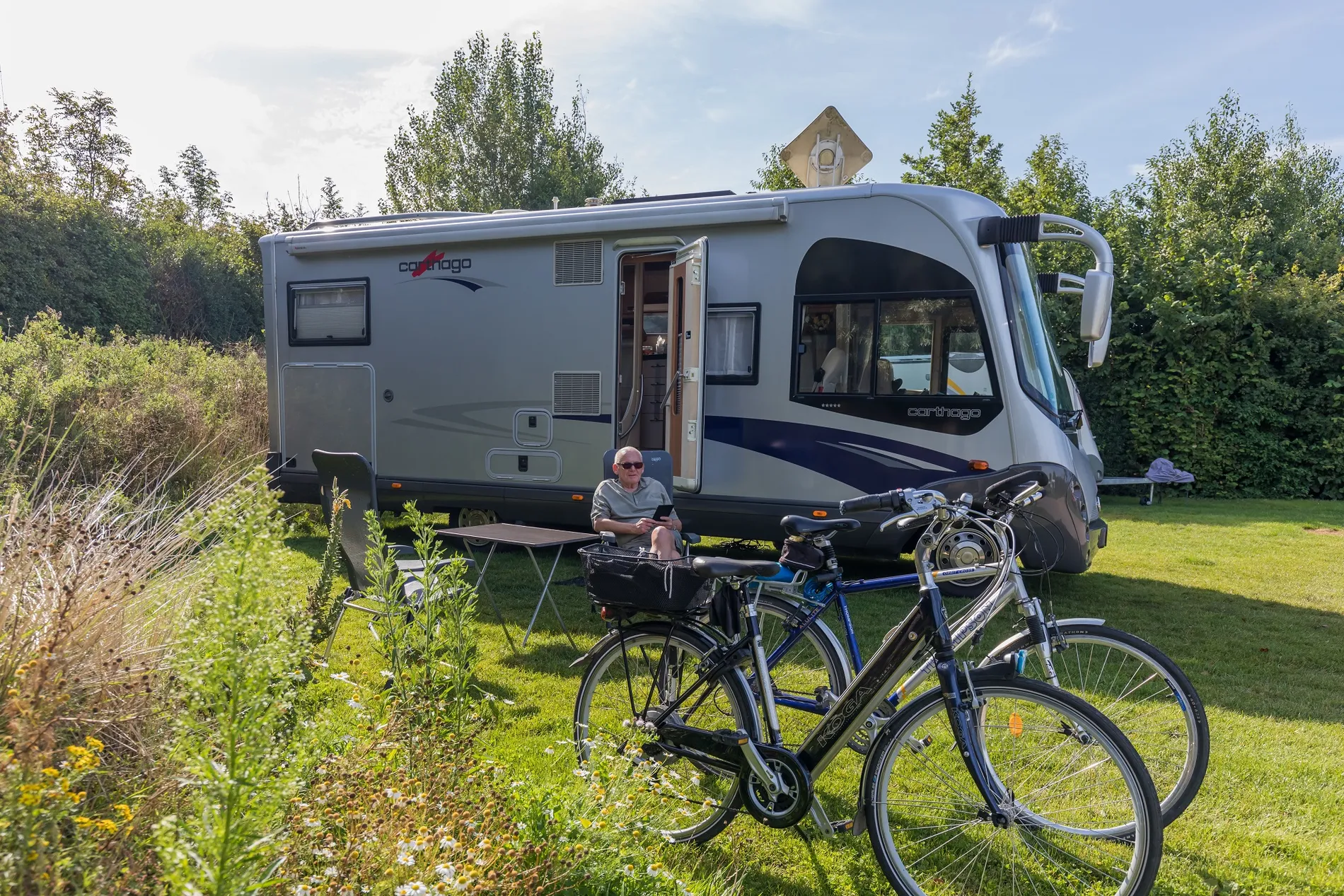 Luxe camperplaats bij Resort Land & Zee met een grote Carthago camper, fietsen en een ontspannen gast in de zon.